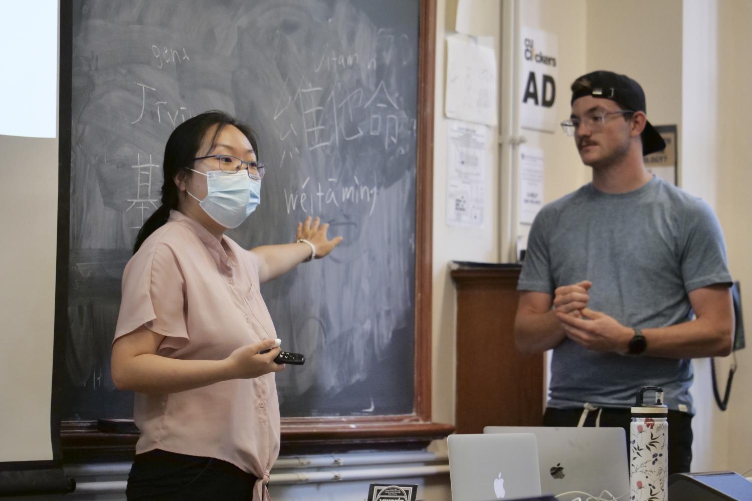 A female professor pointing at a blackboard while a male student is standing next to her