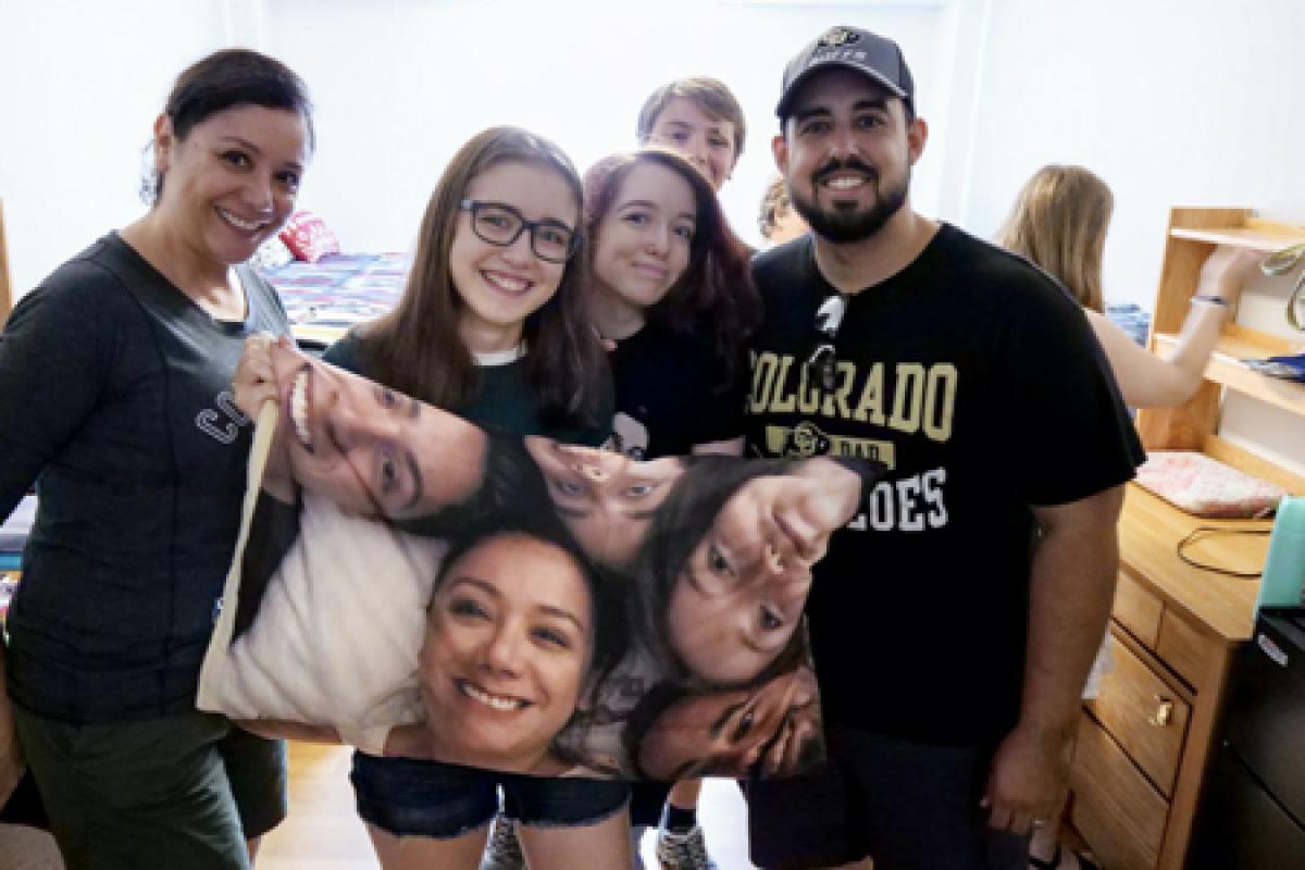 A family poses with their student in a dorm room while holding a keepsake pillowcase