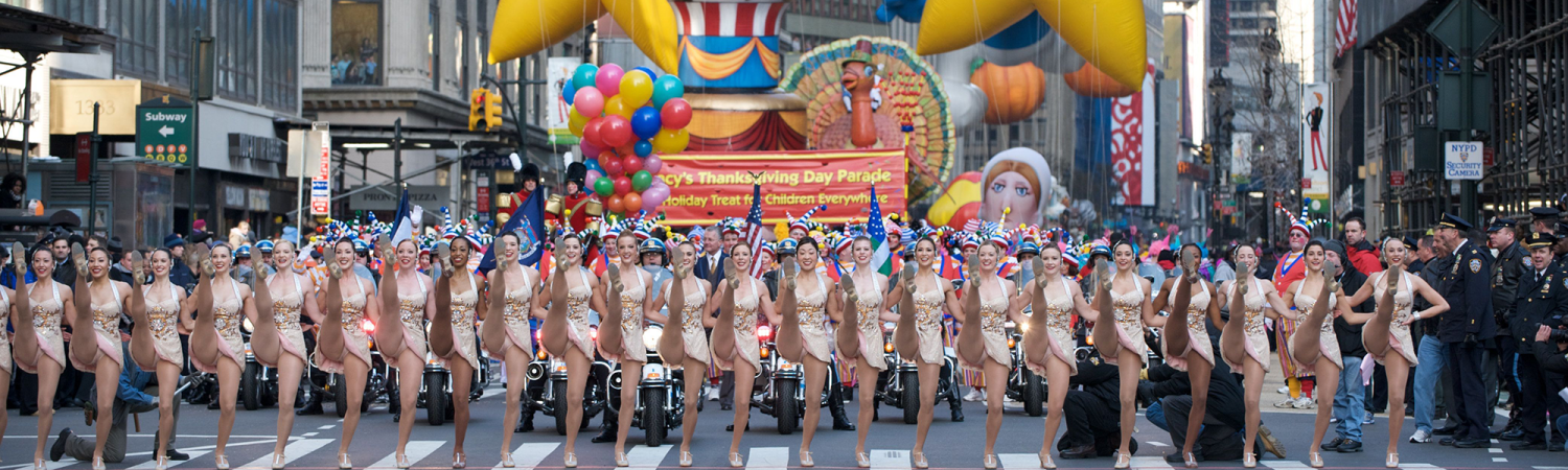 Image of the Macy's Thanksgiving Day Parade with a kick-line of dancers in the foreground and large parade balloons in the background