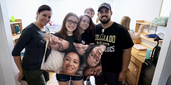 A family poses with their student in a dorm room while holding a keepsake pillowcase