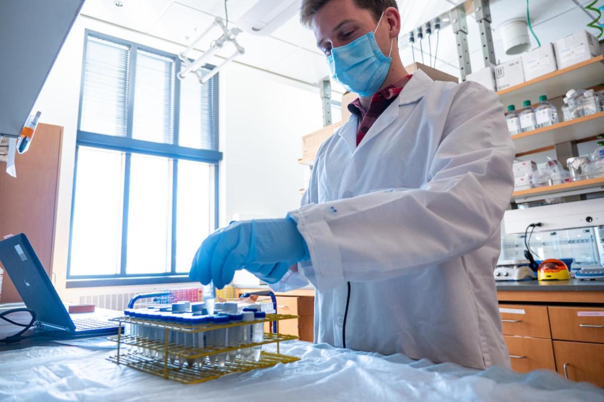 A man wearing a white lab coat and mask analyzes a sample