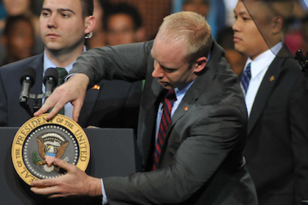 A person wearing a suit adjusts a sign on a lectern that says "President of the United States"