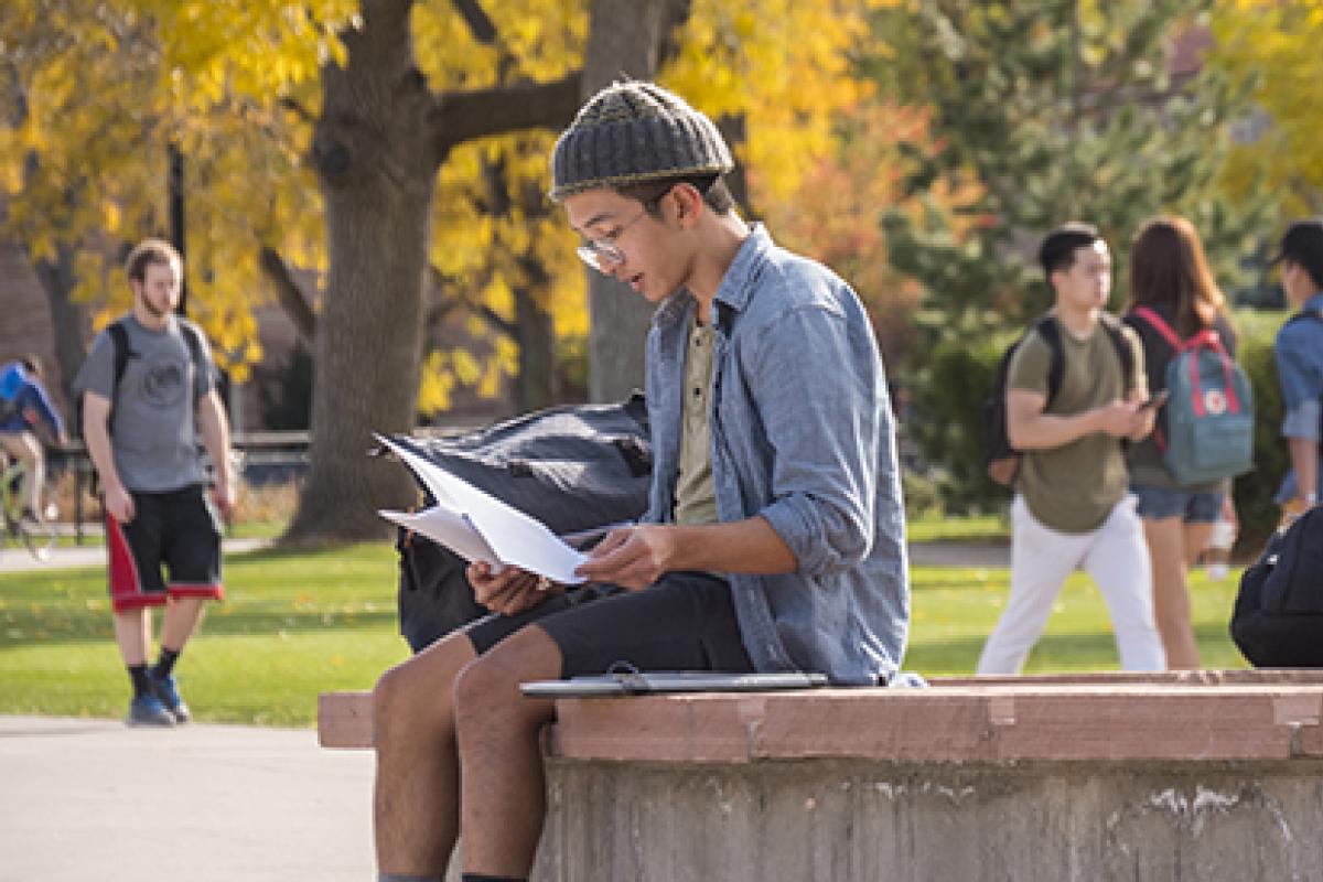A student sits on a concrete column, reading a paper.