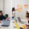 A person casually dressed gestures to a series of sticky notes that seem collected into four groups. The varied dress and seemingly expensive fabrics of four fellow attendees appears to communicate this is a barnstorming session in a tech startup.