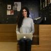 A woman with long, curly hair and a wide smile sits in a fashionable, built-in wood corner. With a laptop on her lap and a space-themed wallpaper filling the background, she appears cheerful at her realized capacity to convey the career value of a liberal arts education.