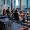 Several people sit in a community workspace housed in a skyrise, looking out over a city.