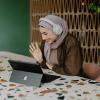 A woman wearing headphones smiles and waves at a computer.