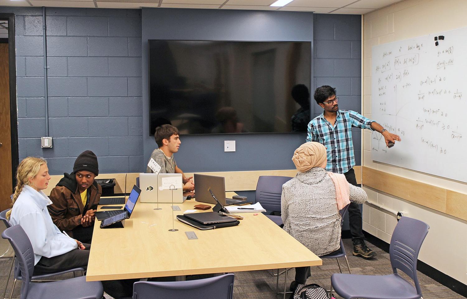 tutor pointing to equation on whiteboard while several students sit at table