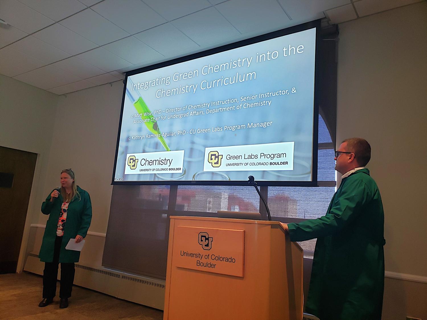 two people in green lab coats in front of screen, presenting about green chemistry