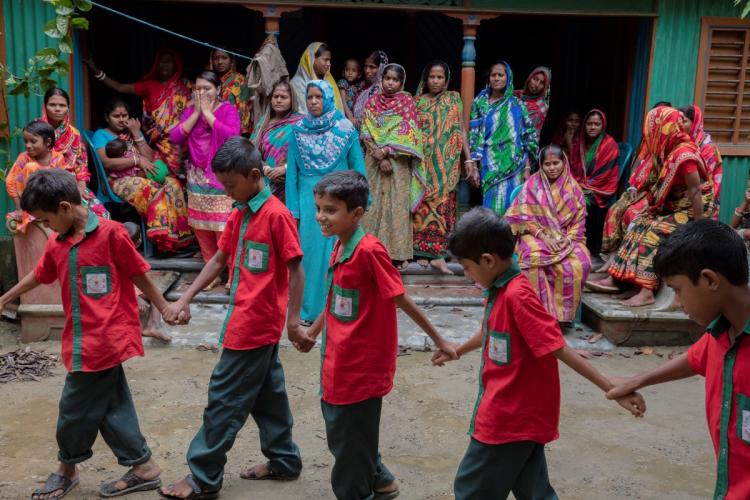 Children in red shirts participating in school activity in Bangladesh with women in saris watching