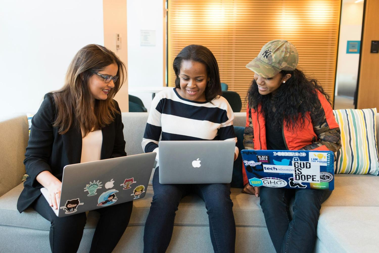 Three smiling women, one white and two Black, sitting on a sofa looking at laptops.