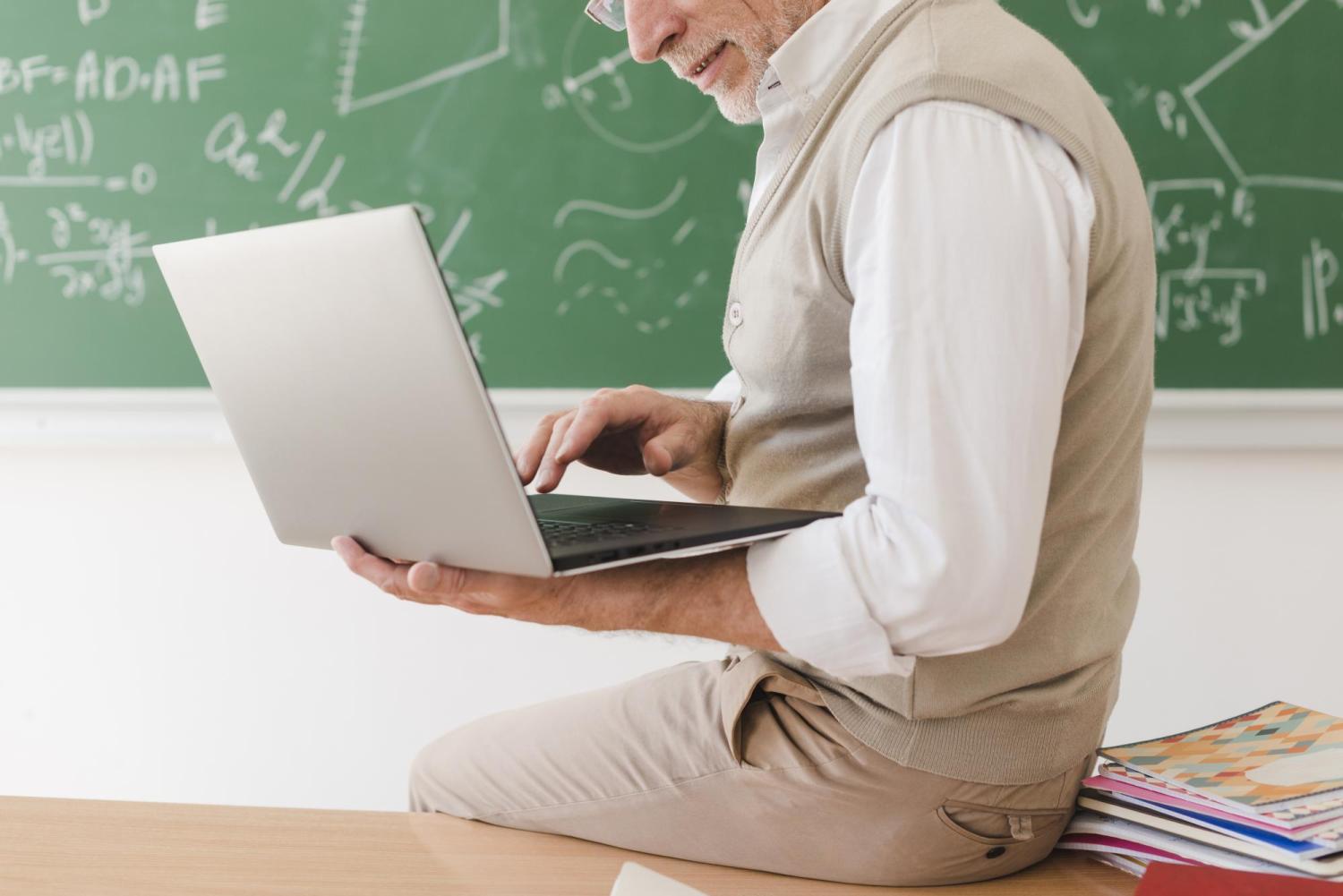 Teacher working on a laptop in front of a chalkboard with written equations