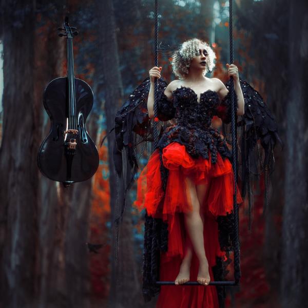 A woman in elaborate black and red garb with wings stands on a wooden swing as a black cello hovers to her right