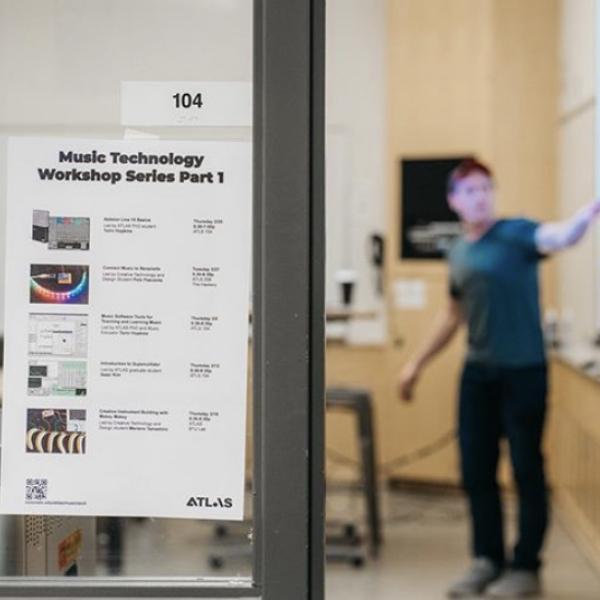 Photo of Music Technology Workshop Series poster hung on wall in front of teacher pointing at screen