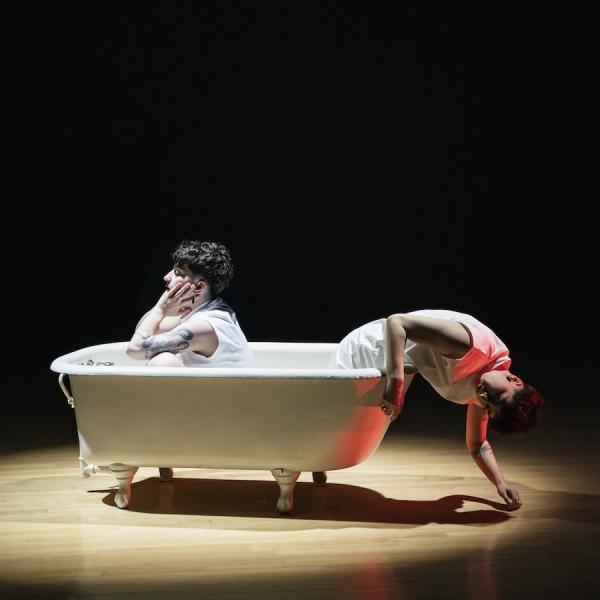 Two people sit languidly in an old-fashioned bathtub on a wood stage with moody lighting