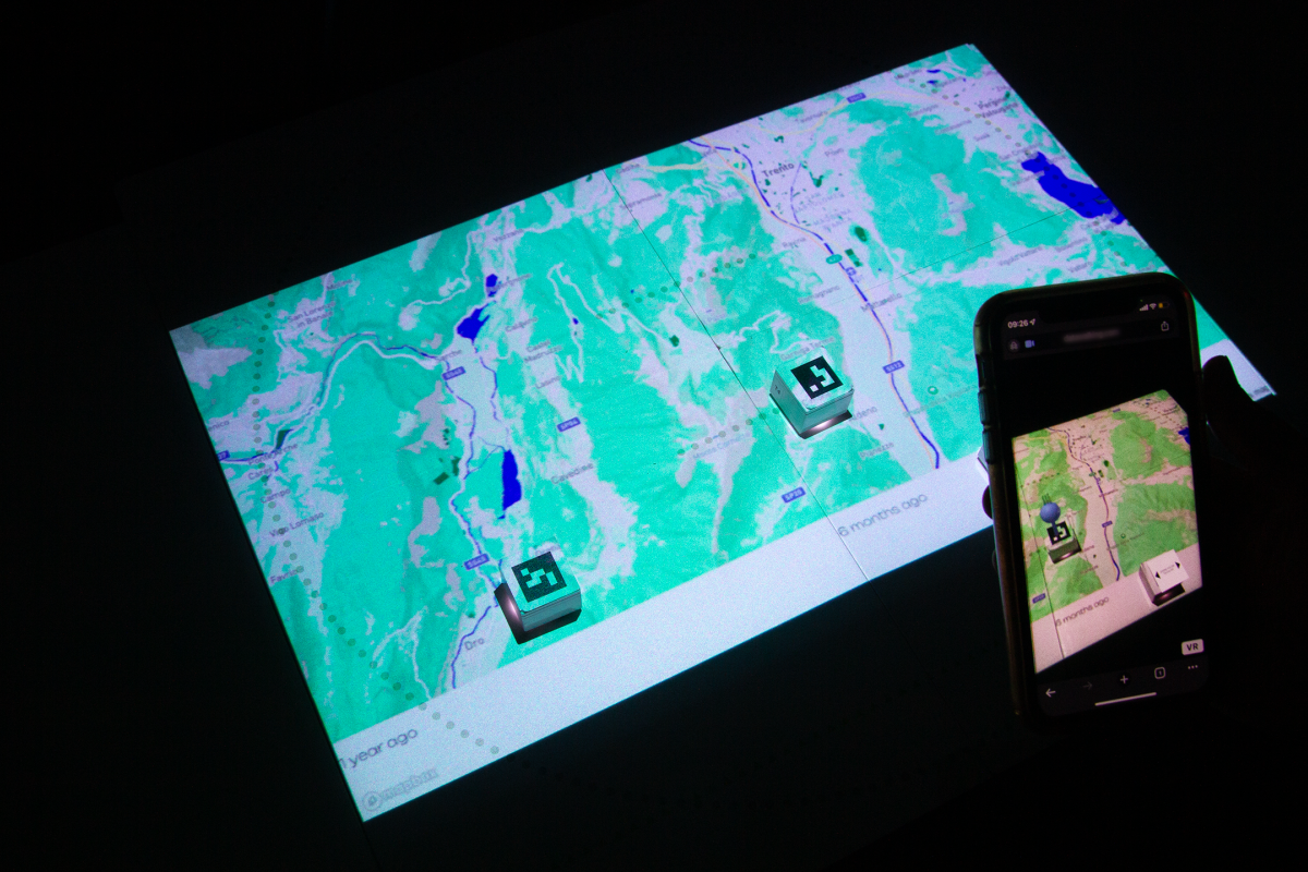 A map projected onto a surface. Two paper blocks are on the map, which show as pins through a phone screen.