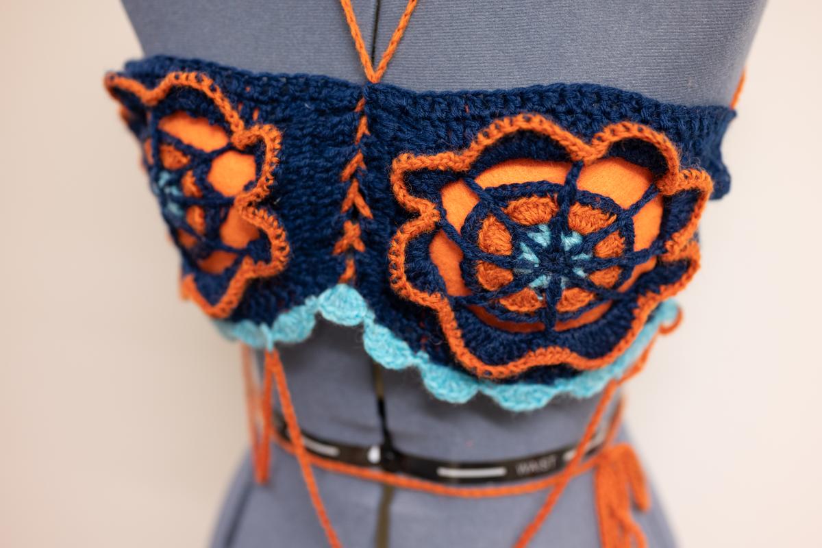 A orange and blue crocheted bralette with a flower designs in the center on a cloth mannequin.