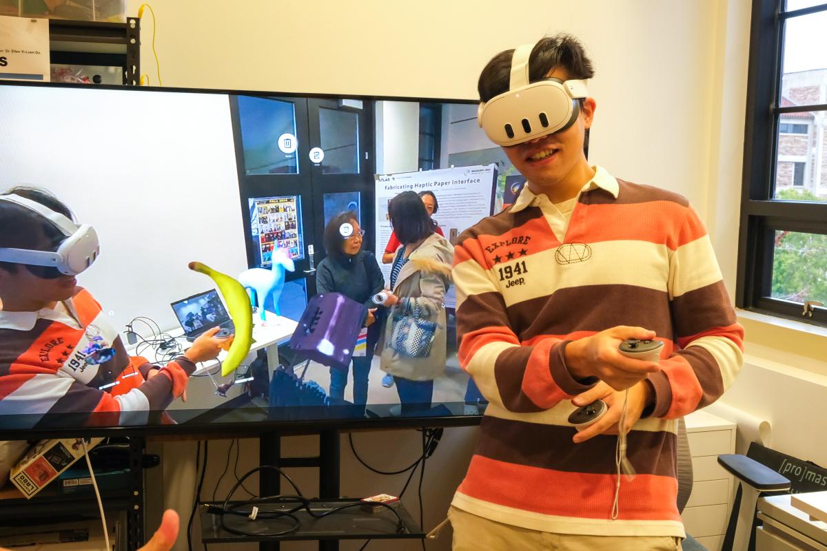 A man stands in front of a TV screen wearing a VR headset. In the TV screen, he is visible interacting with a 3d model of a banana.