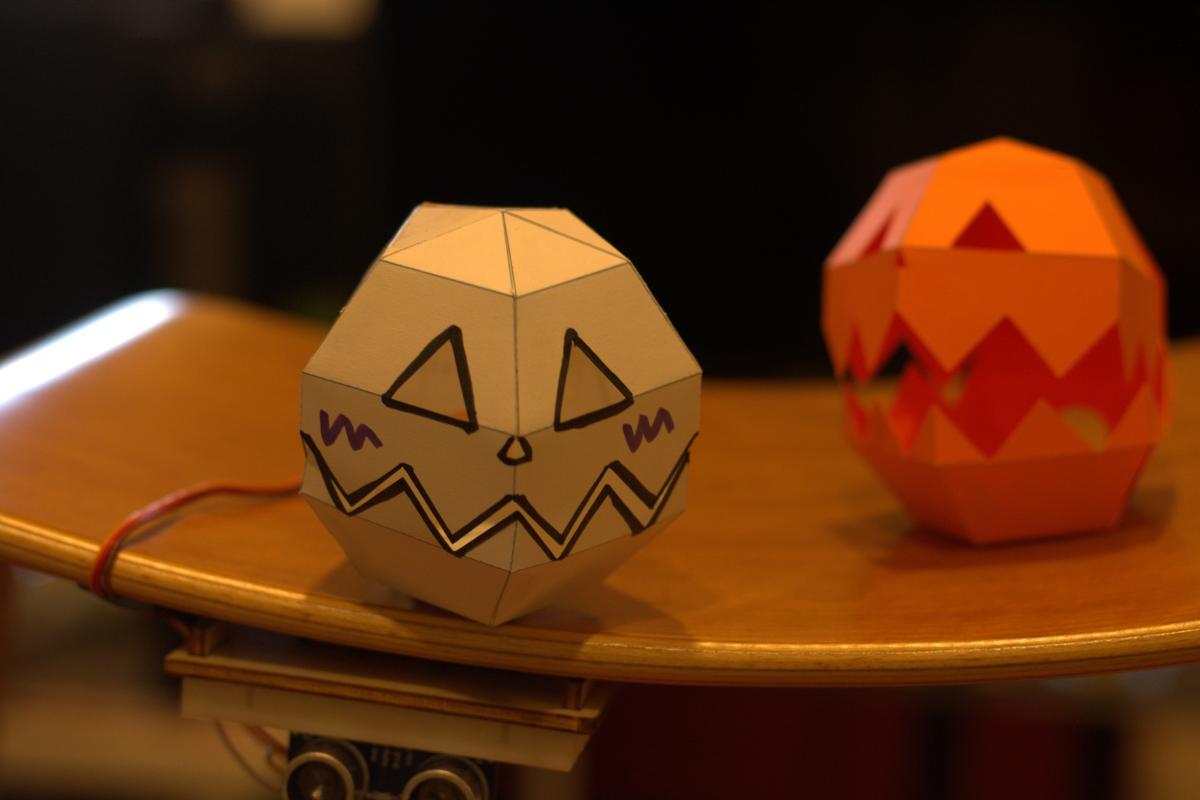Two paper robots shaped like Jacko lanterns sit on a table. One is white and one is orange.