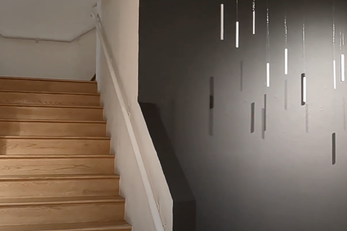 A set of stairs next to a gray wall. The gray wall is lit with a spotlight and features several metal cylinders hanging from the ceiling.