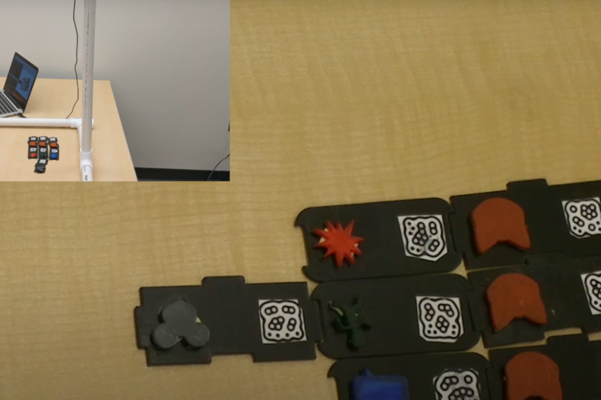On a table are a few black interconnected black pieces with patterns on them. A small screen in the top left corner shows a person on a laptop next to the items.
