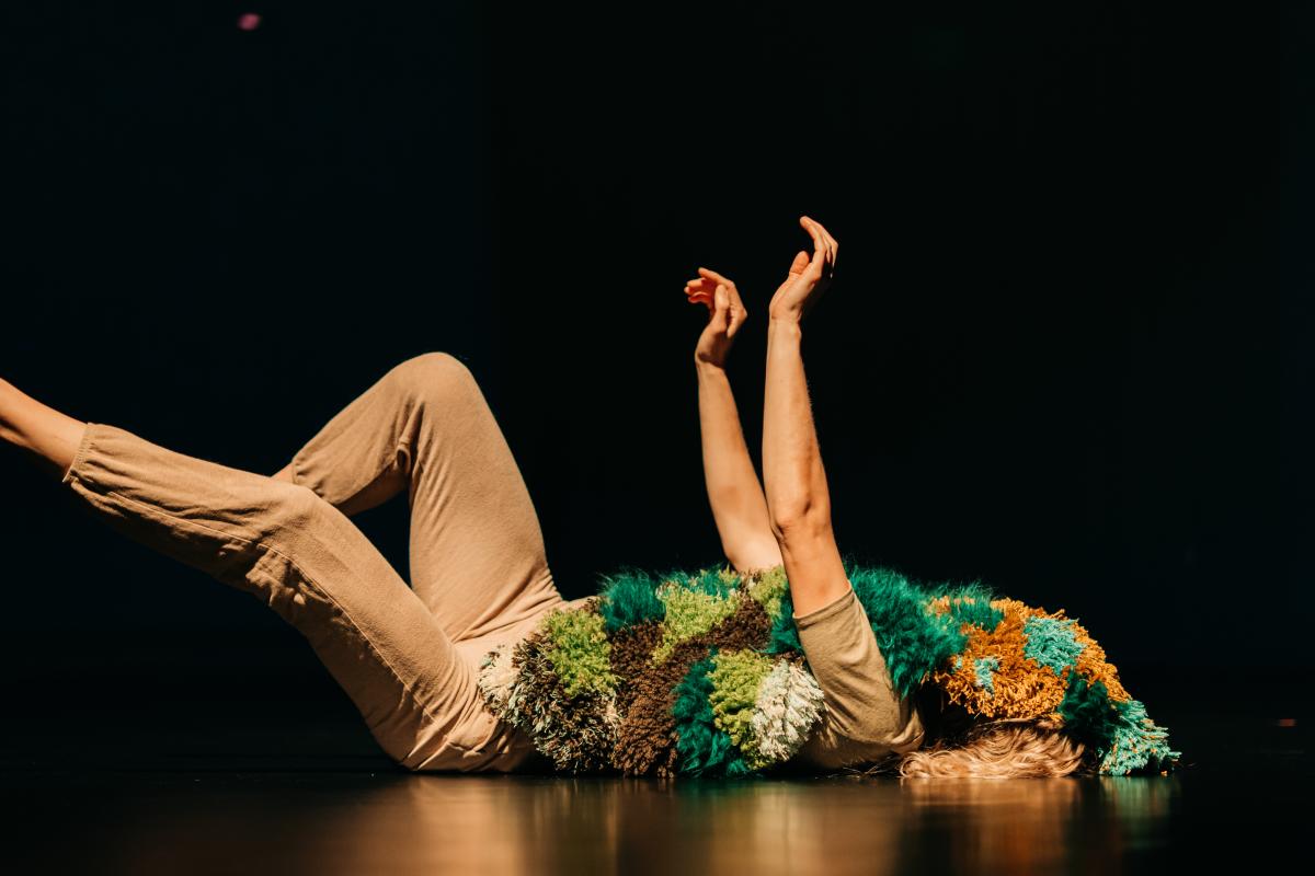 A person in a dark room lays on the floor, arms and legs up in the air. Their torso and face is covered by a rug resembling moss.