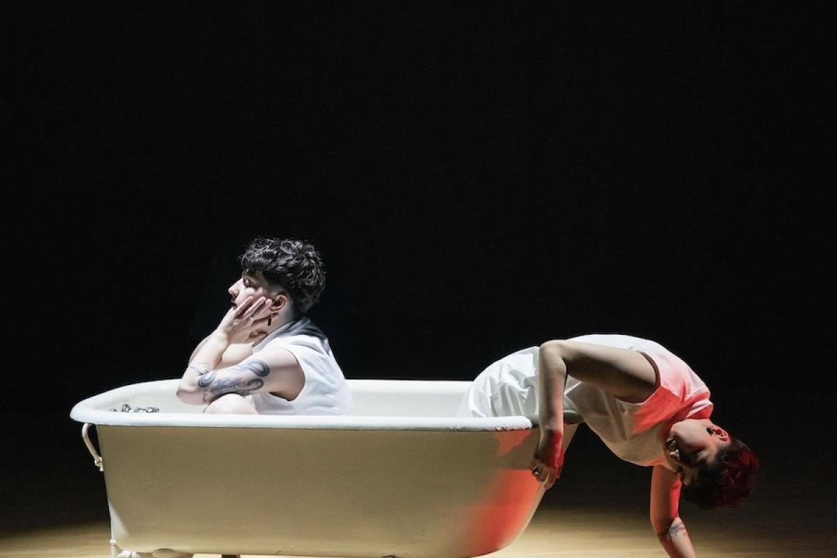 Two people sit languidly in an old-fashioned bathtub on a wood stage with moody lighting