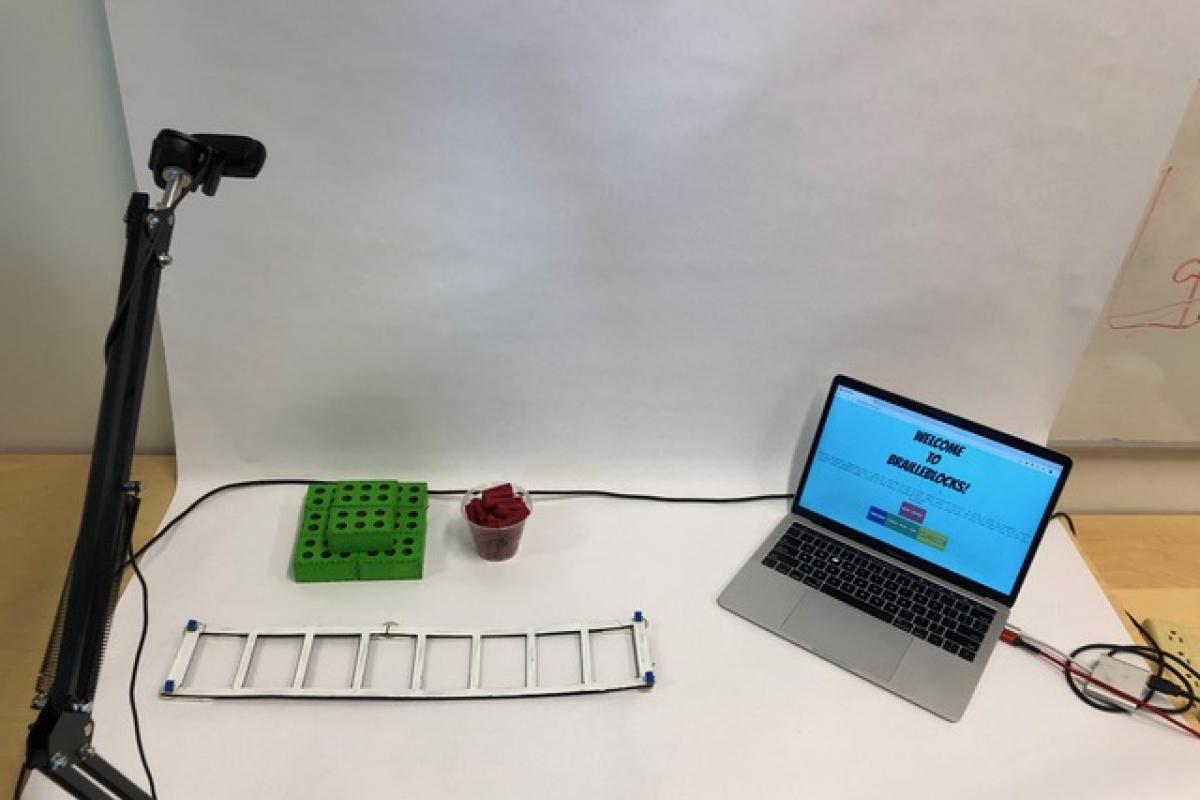 The three components of the BrailleBlocks system on a table. On the left is a webcam which points downwards onto the table. In the middle are stacked green blocks which resemble Braille cells. Next to the blocks is a frame with rectangle slots where the blocks will be placed. On the right is a laptop showing the home screen of the BrailleBlocks web application.