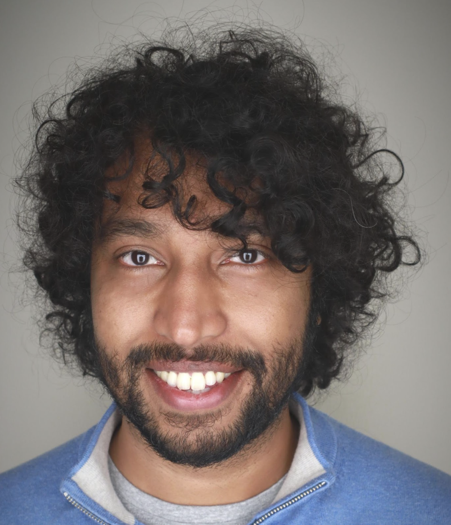 A man with brown skin, curly black hair and a short beard smiles at the camera.