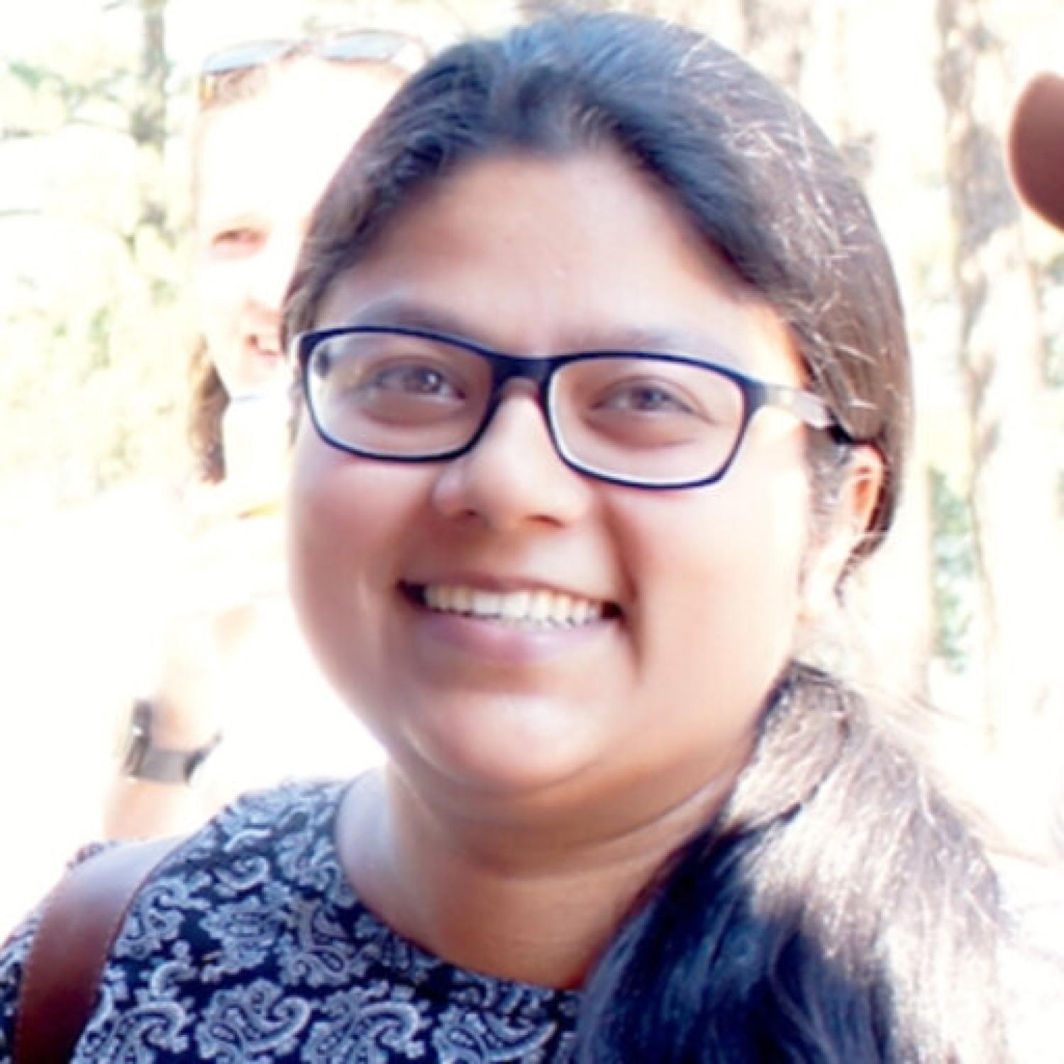 Profile image of a brown woman with long black hair in a pony tail and glasses, smiling.