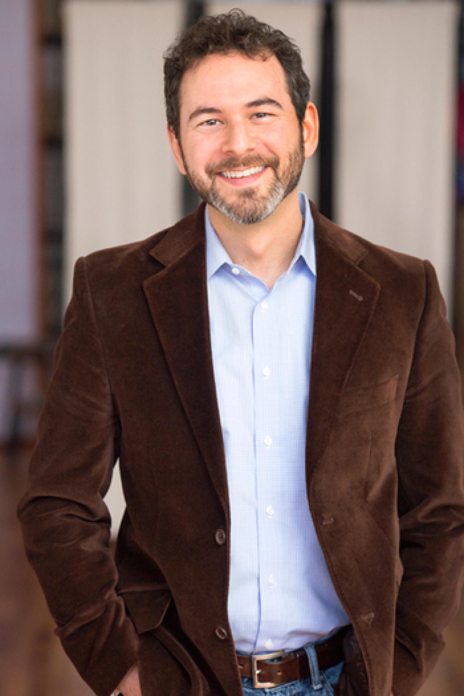 Half-body of a white man with short black hair and beard. He is wearing a brown and periwinkle suit.