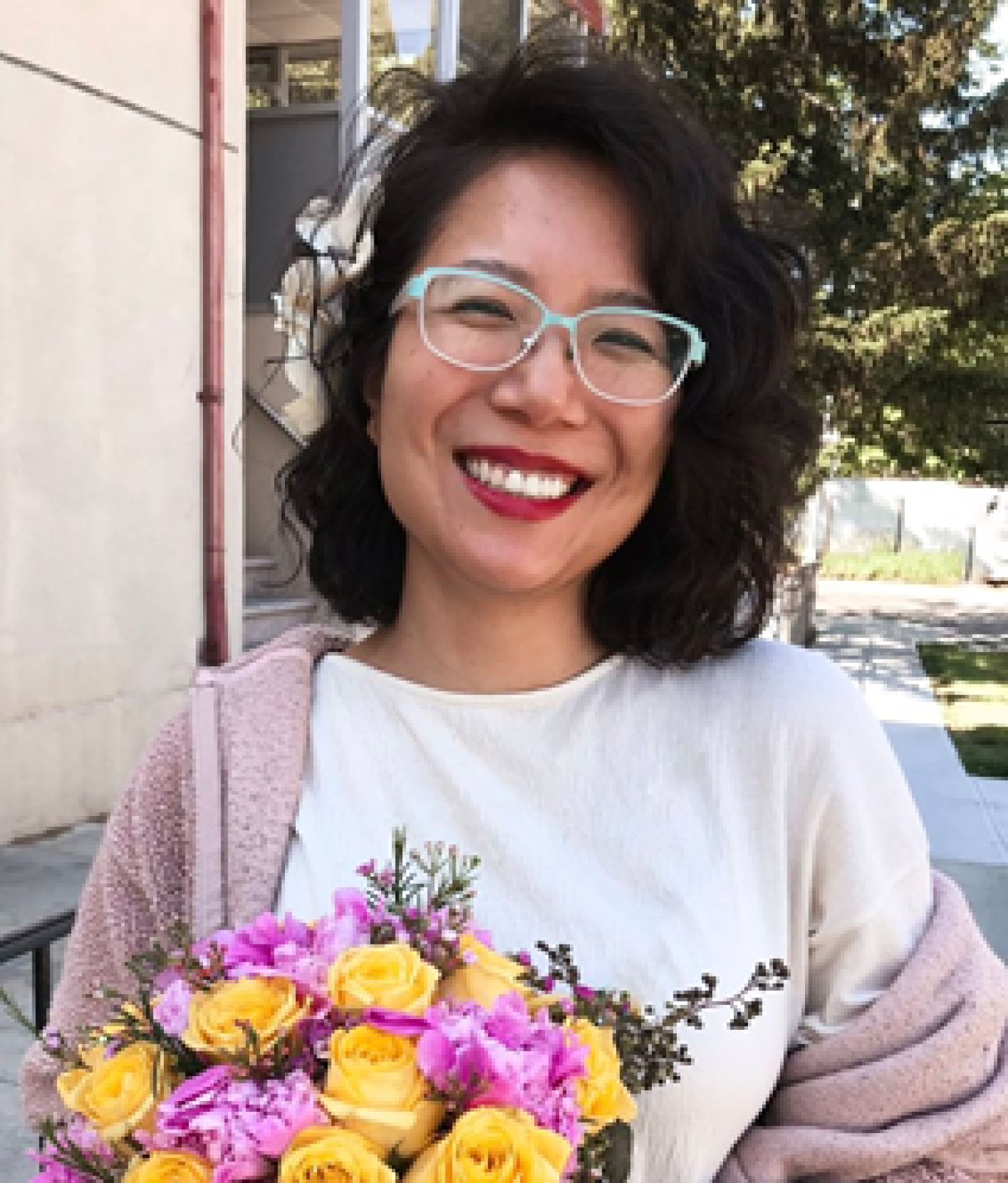 Profile image of an Asian woman with a curly black bob smiling. She is wearing glasses and holding a bouquet of flowers.