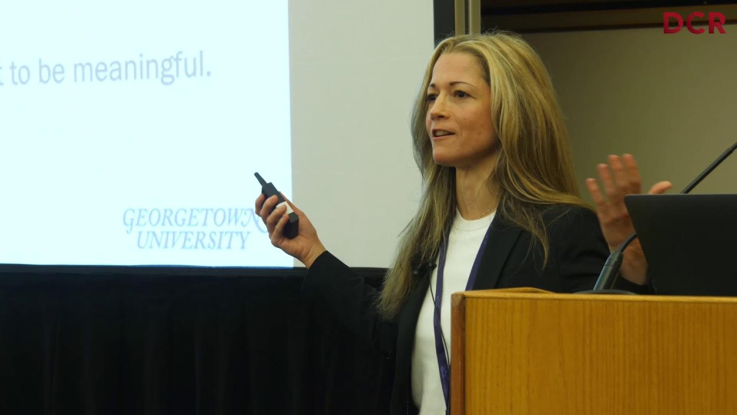 White woman with long blonde hair, speaking on stage in front of a podium.