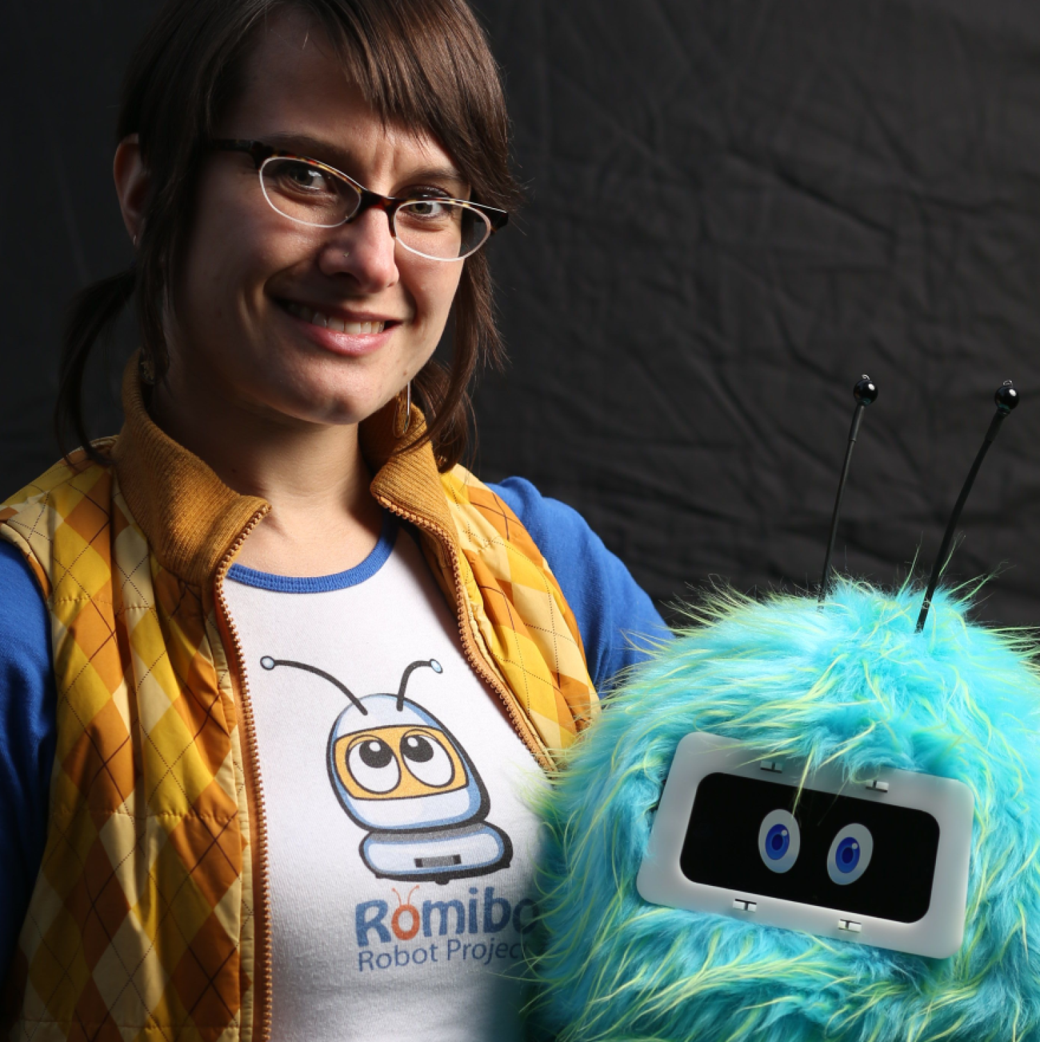 Half-body of a white woman with brown hair tied back and glasses. She is holding a blue fuzzy robot.