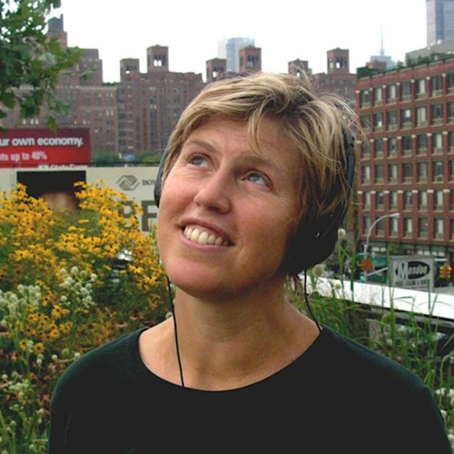 Profile of a middle-aged white woman looking up, smiling. She has short blonde hair and is wearing headphones.