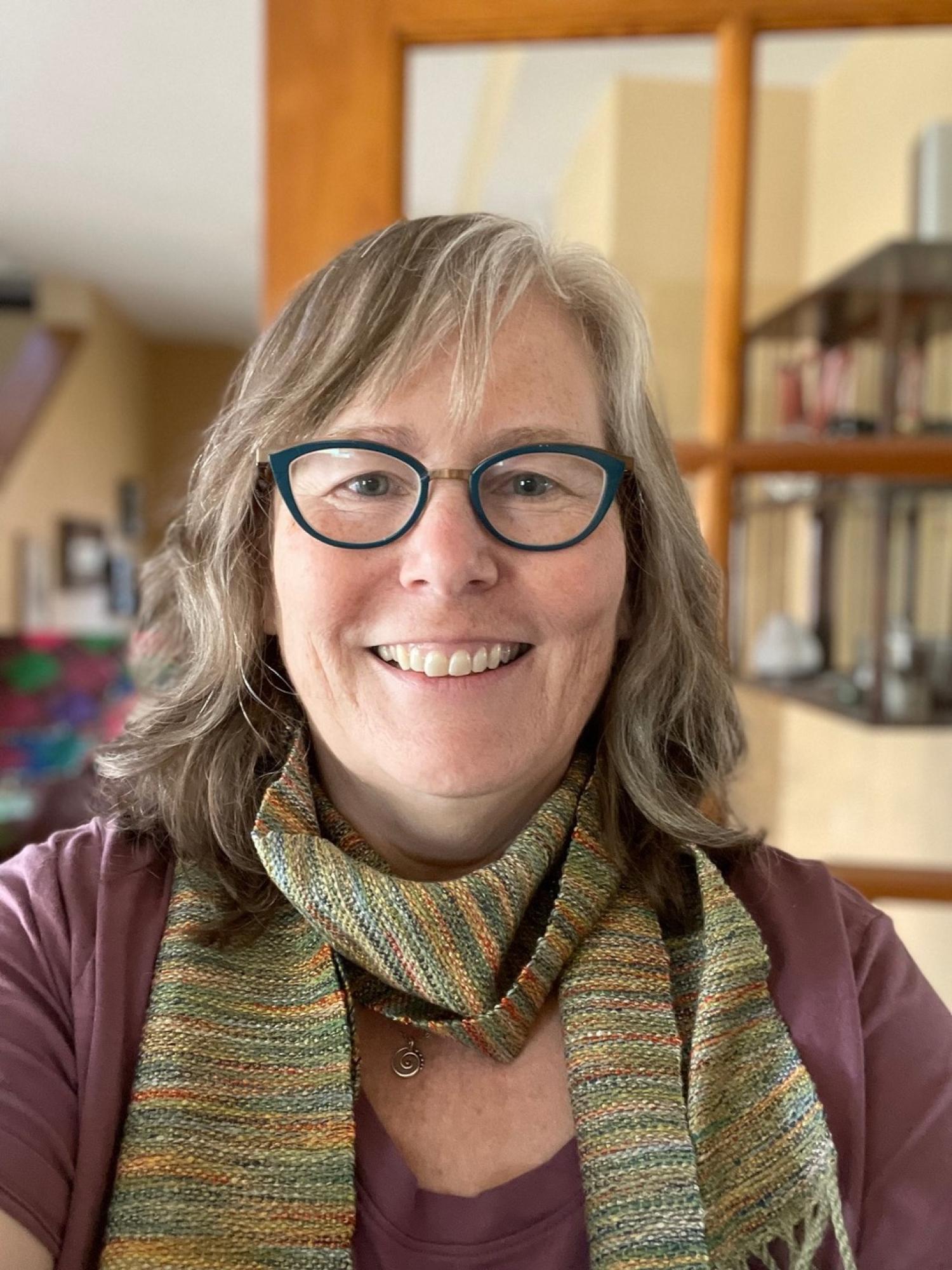 Profile of an older white woman with medium length gray hair and glasses, smiling.
