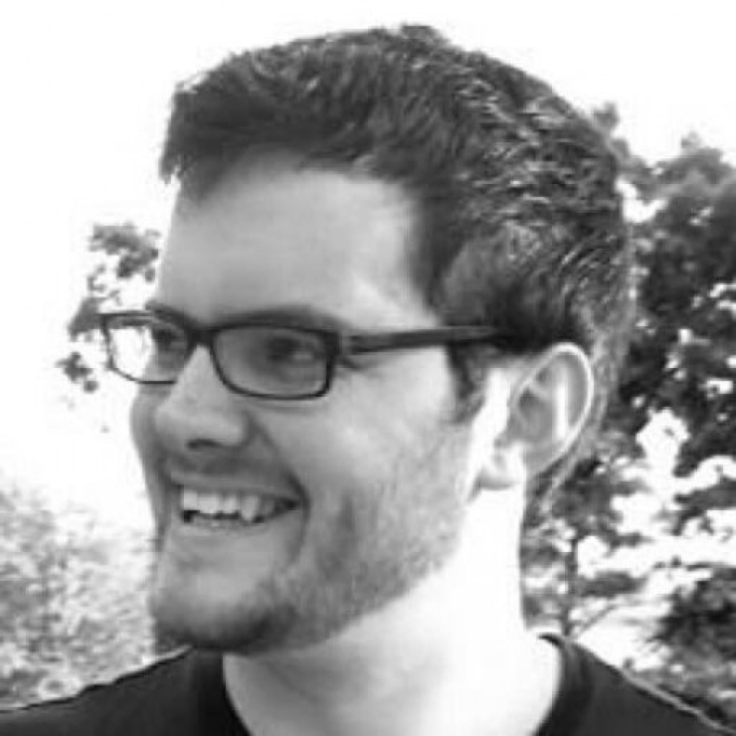 Black and white profile of a white man with short dark hair, stubble and glasses.