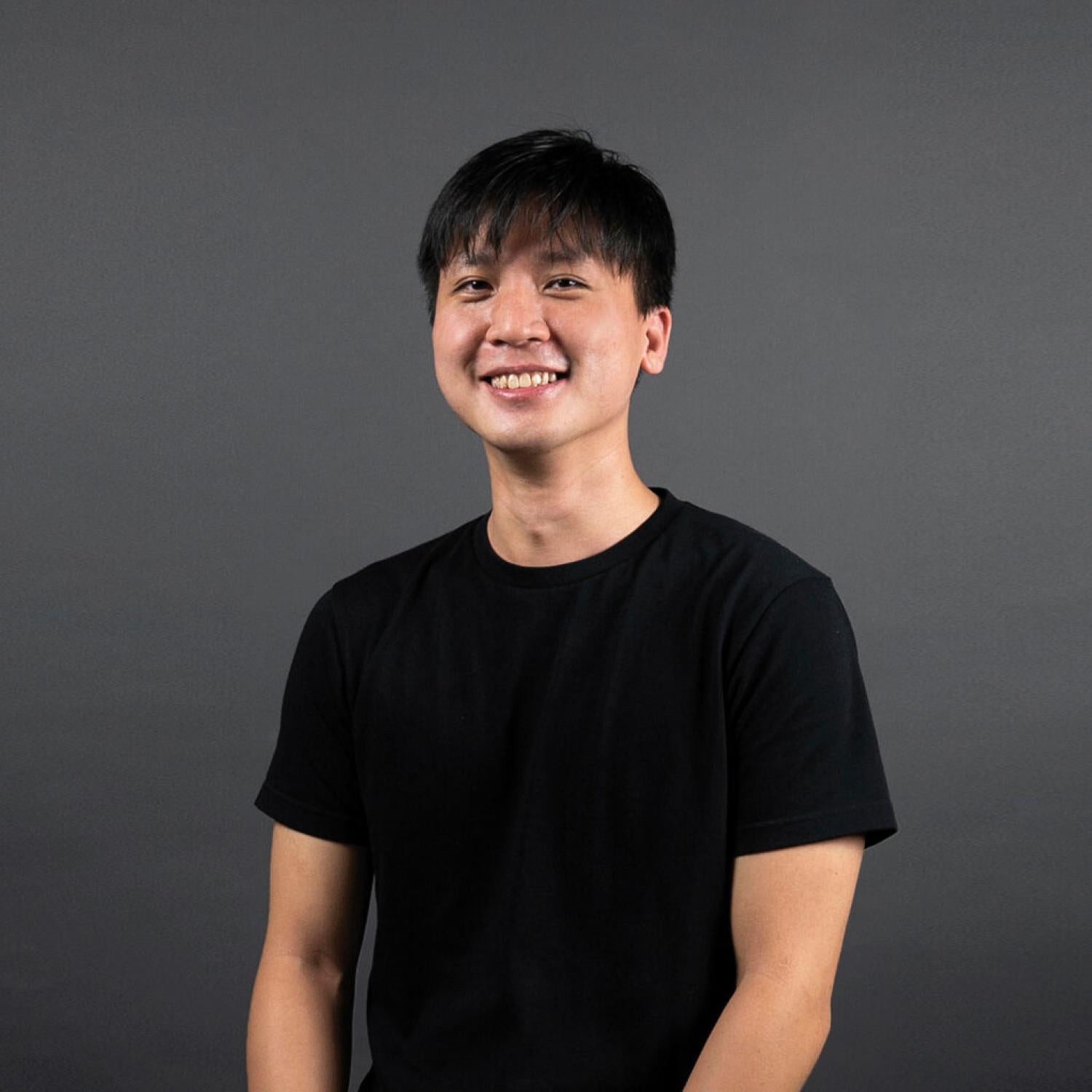 Half-body of a young asian man with short black hair, smiling. He is wearing a black t-shirt.
