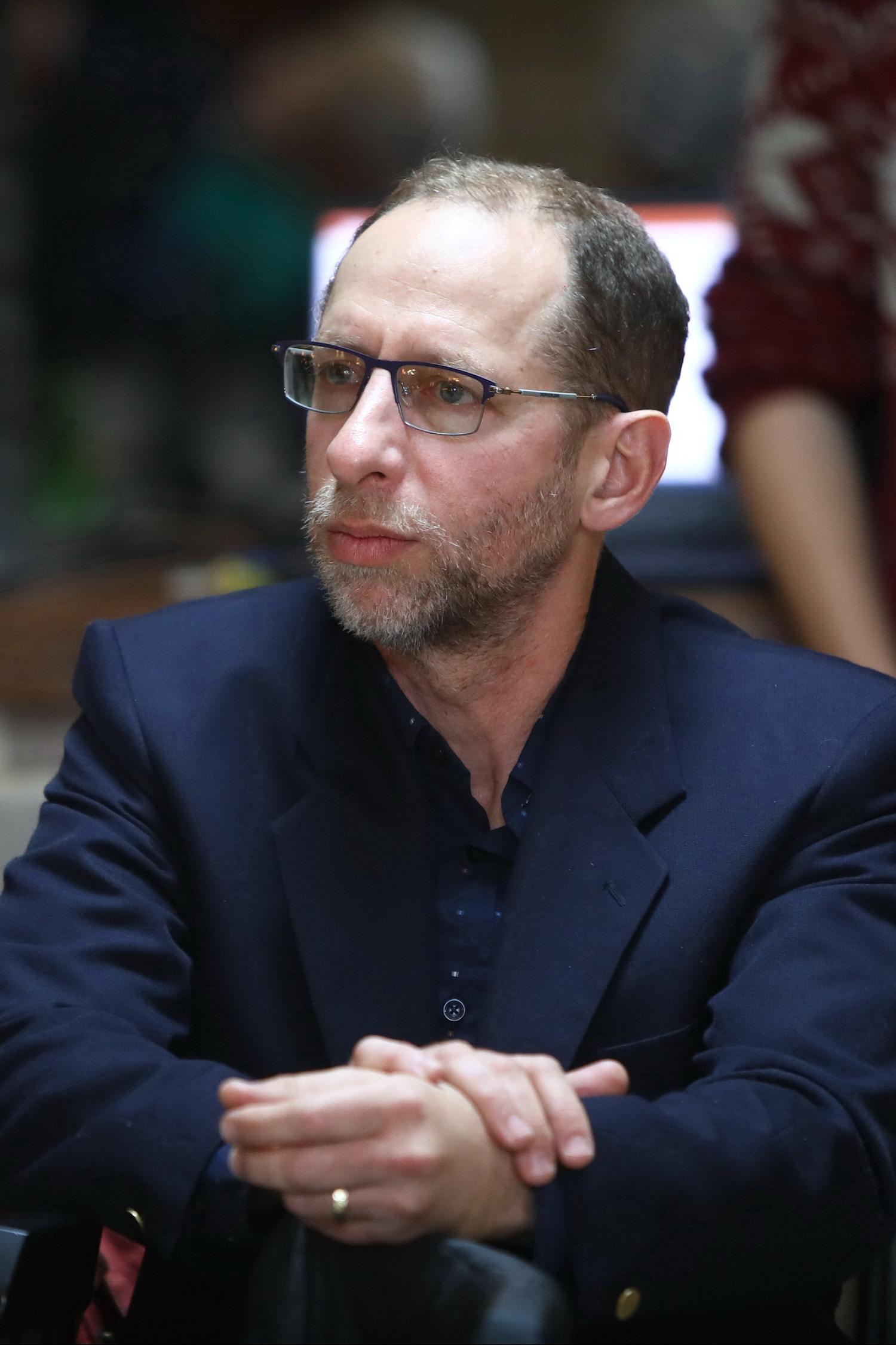 Waist-up of an older white man with glasses and short gray hair and beard. His hands are clasped in front of him and he is wearing a navy suit jacket.