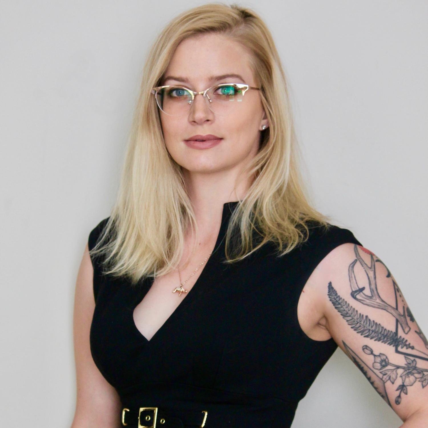 Waist-up image of a young white woman with long blonde hair. She is wearing glasses and has visible tattoos on her arm.