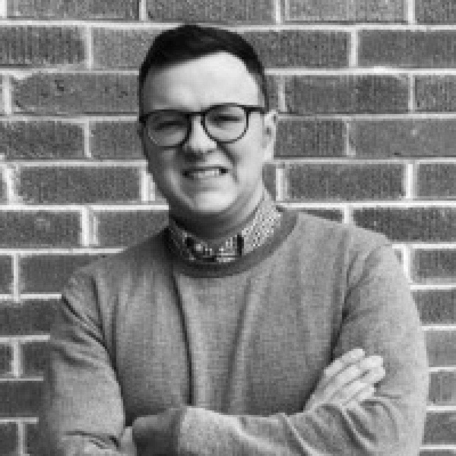 Profile of a white man with short dark hair in black and white. He is wearing glasses and a sweater.