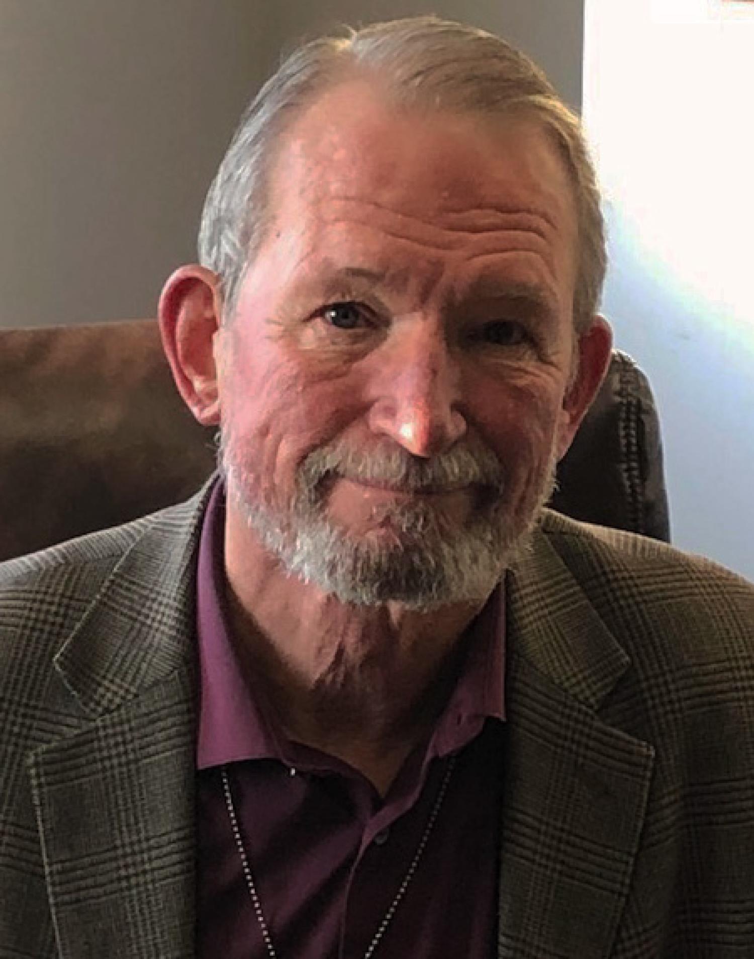 Profile of a older white man with short gray hair and beard, smiling. He is wearing a brown and burgundy suit.