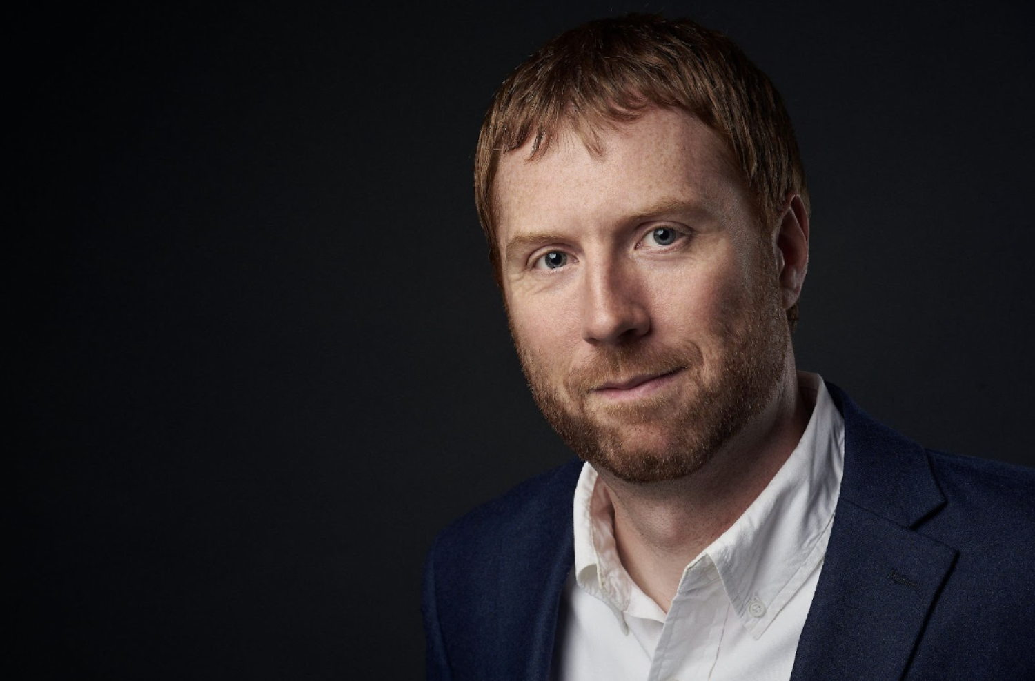 Profile of a white man with short red hair and beard. He is wearing a black suit.