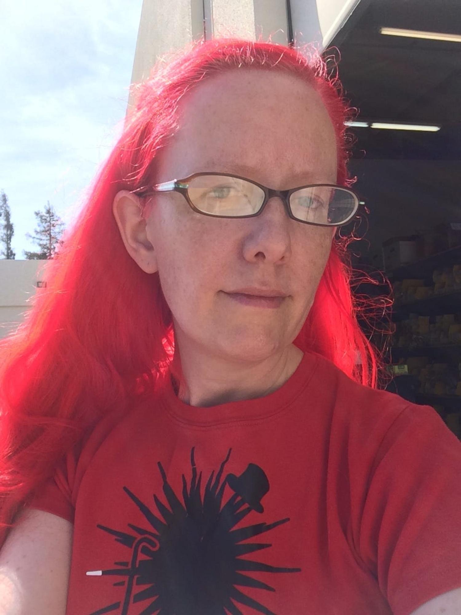 Profile of an older white woman with long scarlet hair. She is wearing glasses and a red t-shirt.