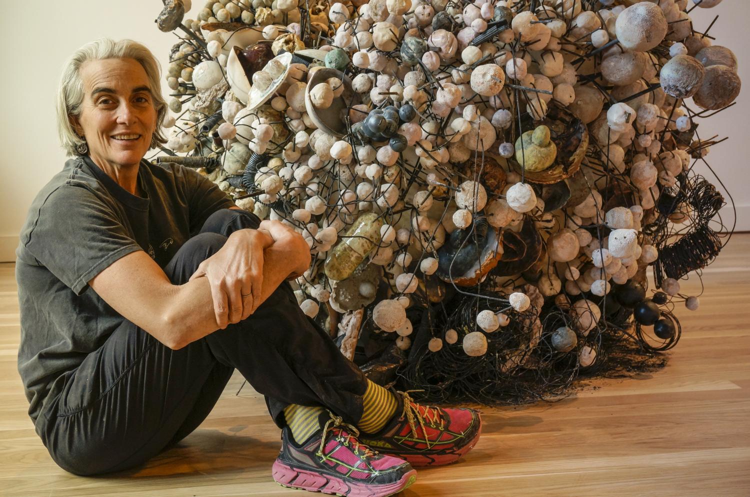 Older woman siting on the floor hugging her knees next to a complex art sculpture.