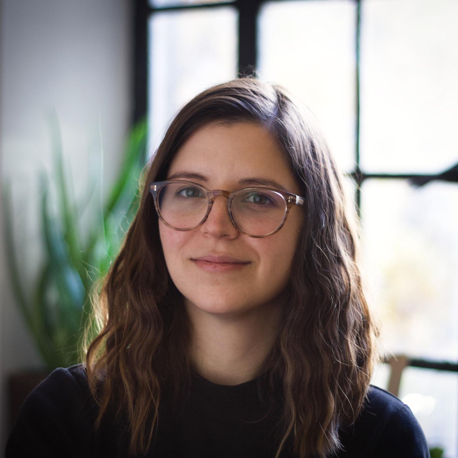Profile of a white woman with long wavy brown hair. She is wearing a black shirt and glasses.