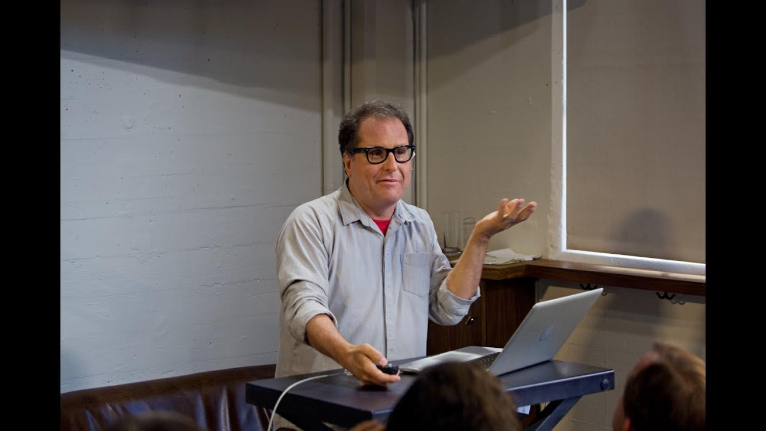 A half-body of an older white man standing at a podium, speaking. He is wearing glasses and a blue shirt.