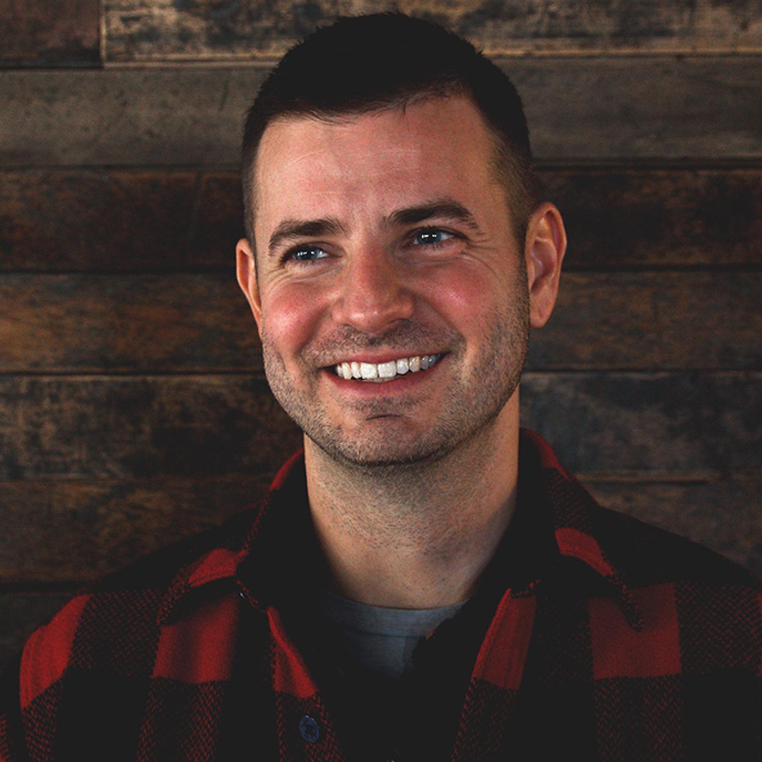 Profile of a white man with short black hair and stubble. He is wearing a red flannel.
