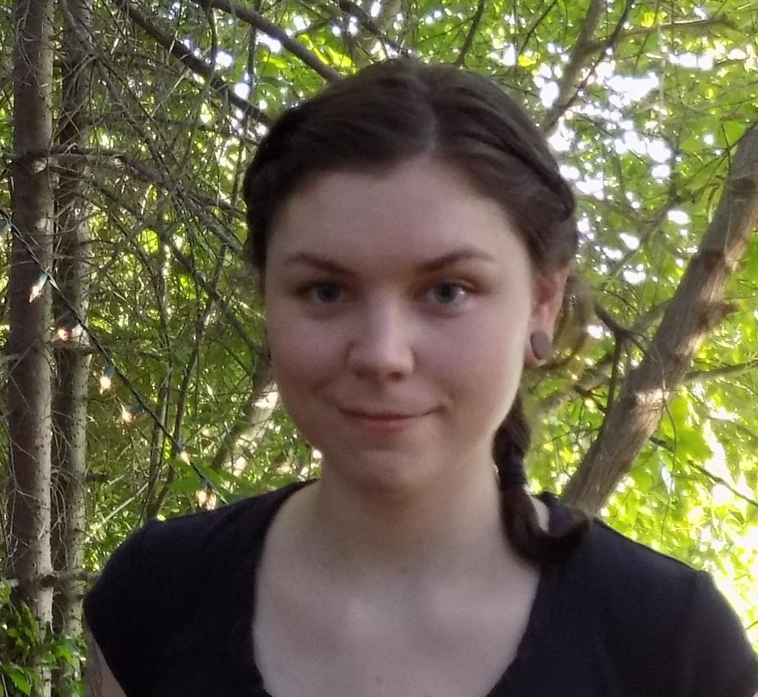 Profile of a young white woman in a forest with dark brown hair tied back, smiling.