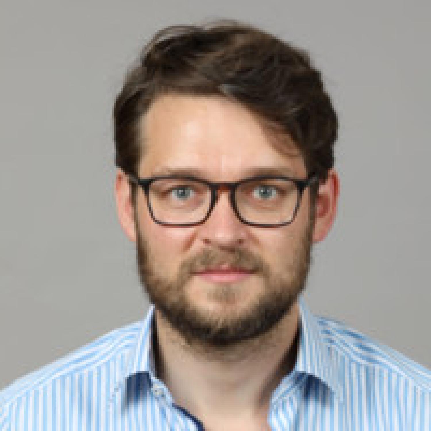 Profile of a white man with short brown hair and a beard. He is wearing glasses and a blue dress shirt.